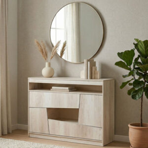 Cómoda Kendor de melamina color madera clara Helsinki, con diseño de cajones asimétricos y un estante abierto, ambientada en un dormitorio de tonos neutros con un espejo circular y una planta de interior.