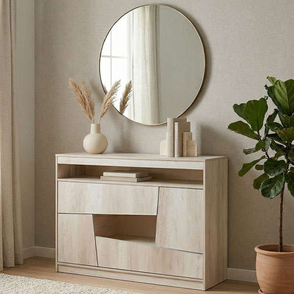 Cómoda Kendor de melamina color madera clara Helsinki, con diseño de cajones asimétricos y un estante abierto, ambientada en un dormitorio de tonos neutros con un espejo circular y una planta de interior.