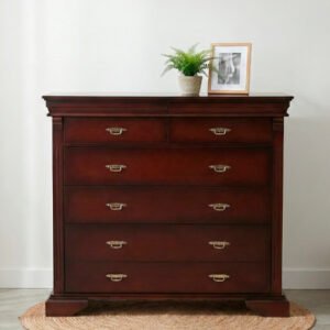 Cómoda de madera estilo Versalles en acabado cedro sombreado con cinco cajones, herrajes de bronce y detalles tallados, ubicada en un ambiente minimalista con piso de cemento y pared blanca.