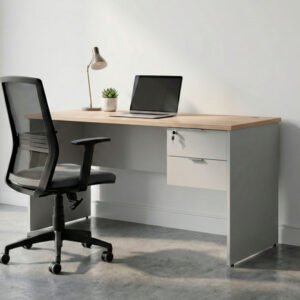 Escritorio de oficina moderno con superficie de madera clara, estructura ceniza, pasacables y cajonera lateral de dos cajones con llave, ubicado sobre un piso de cemento alisado frente a una pared blanca.