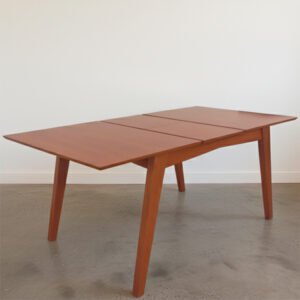 Mesa de comedor rectangular extensible modelo Andrea, fabricada en madera de paraíso con acabado natural, mostrando su sistema de apertura central sobre un piso de cemento alisado y fondo blanco.