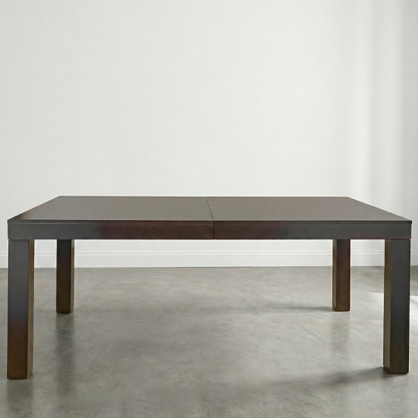 Mesa de comedor rectangular modelo Mónaco, extensible y de color marrón oscuro, con patas gruesas y diseño minimalista sobre suelo de cemento