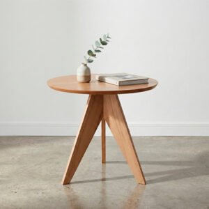 Mesa de centro redonda modelo Aren de madera clara con patas estilo trípode, ambientada en un living moderno con piso de cemento alisado, pared blanca y decoración minimalista con lámpara de pie.