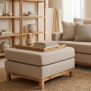 Un plano medio de un puff otomana cuadrado de la colección Bambu, tapizado en tela texturizada beige y con patas de madera clara, posado sobre una alfombra de yute. Encima del puff, hay una bandeja de bambú con libros y una vela. Al fondo, se ve parte del sofá Bambu coordinado, una lámpara de pie trípode de madera y estanterías de madera con cerámica artesanal.
