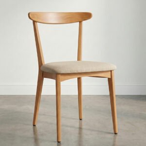 Silla de comedor de madera clara con respaldo curvo y asiento tapizado en tela color arena, situada en una habitación con suelo de cemento pulido y pared blanca con zócalo.