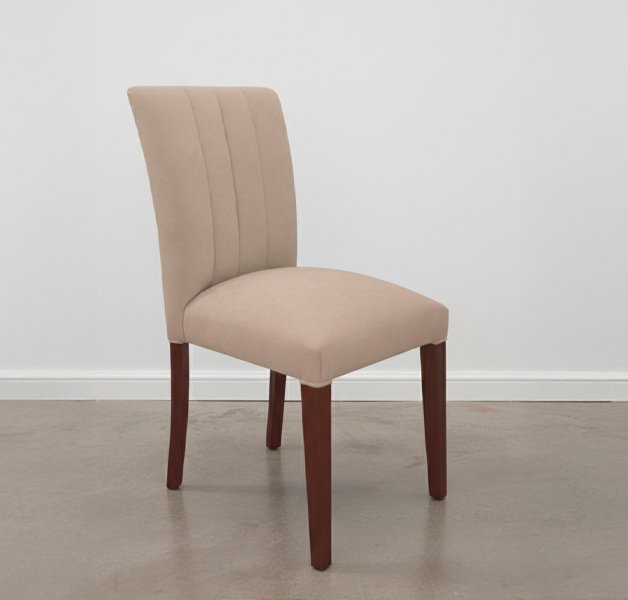 Silla tapizada en color beige con patas de madera oscura, ubicada en una habitación con piso de microcemento gris, pared blanca con zócalo y luz natural suave.