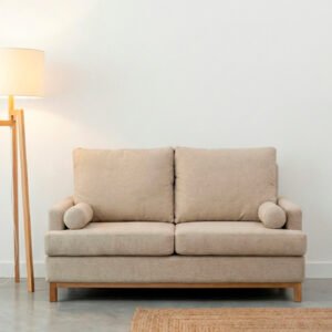 Sofá modelo Bambú de dos cuerpos tapizado en tela color natural, ubicado en una sala moderna con piso de cemento alisado, lámpara de pie y estantería de madera.