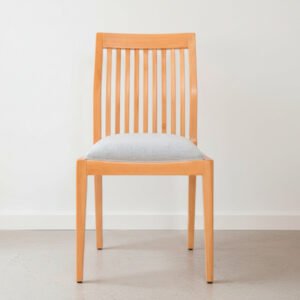 Silla de comedor modelo Olsen Natura de madera clara con respaldo de varillas verticales y asiento tapizado en color gris claro, ubicada sobre un suelo de cemento alisado frente a una pared blanca