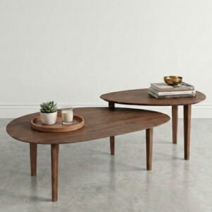 Conjunto de dos mesas de centro anidadas de madera de nogal con formas orgánicas, ubicadas sobre un piso de concreto pulido en una sala minimalista con una pared blanca. La mesa inferior tiene una bandeja de madera con una maceta y una vela, y la mesa superior tiene una pila de libros con un cuenco dorado.