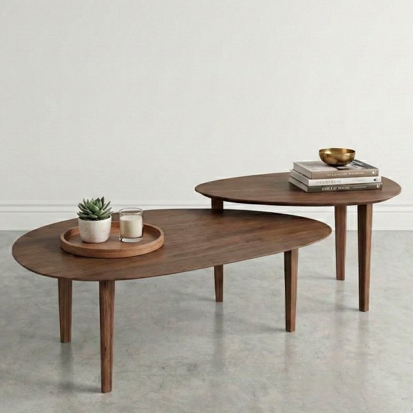 Conjunto de dos mesas de centro anidadas de madera de nogal con formas orgánicas, ubicadas sobre un piso de concreto pulido en una sala minimalista con una pared blanca. La mesa inferior tiene una bandeja de madera con una maceta y una vela, y la mesa superior tiene una pila de libros con un cuenco dorado.