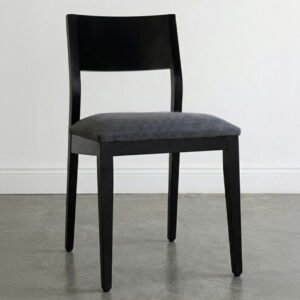 Silla de comedor modelo Humberto fabricada en madera de guatambú teñida de negro. Posee un respaldo cerrado y recto, asiento acolchado tapizado en tela gris oscuro, situada sobre un piso de cemento alisado con iluminación natural suave.