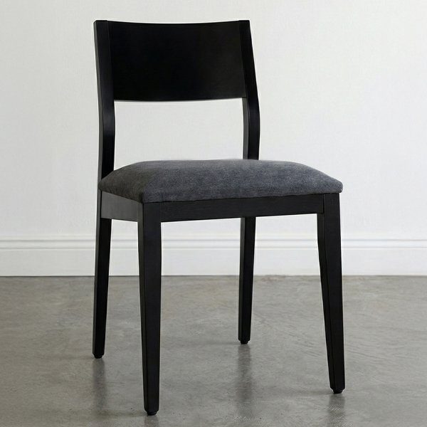 Silla de comedor modelo Humberto fabricada en madera de guatambú teñida de negro. Posee un respaldo cerrado y recto, asiento acolchado tapizado en tela gris oscuro, situada sobre un piso de cemento alisado con iluminación natural suave.