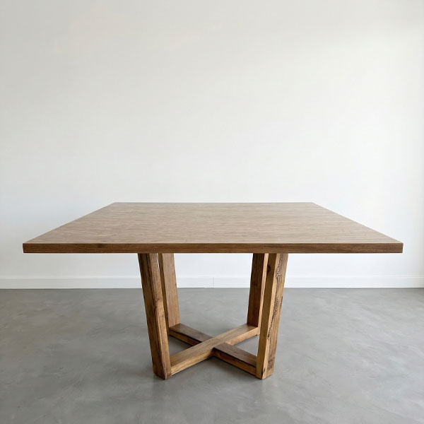 Mesa de comedor cuadrada modelo Ámbar con tapa enchapada en madera de petiribí y base de patas macizas cruzadas, sobre un suelo de cemento alisado en un ambiente minimalista.