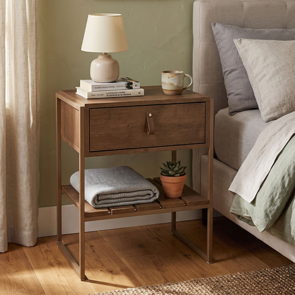 Mesa de Luz James de estilo industrial con cajón de madera y estante inferior, junto a una cama con sábanas grises y verdes.