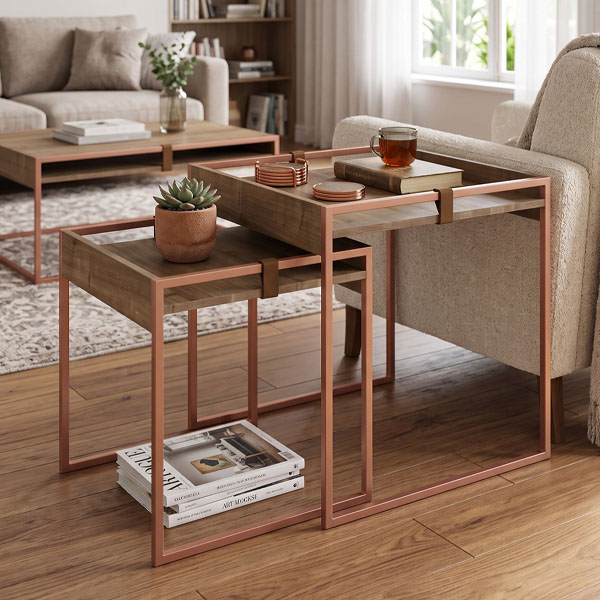 Juego de dos mesas de arrime nido de estilo industrial moderno, con estructura metálica en tono cobre rosado y tapas tray en madera de roble texturizada con detalles de cinchas de cuero. La mesa más alta se ubica junto al apoyabrazos de un sofá de living y sostiene una taza de cerámica.